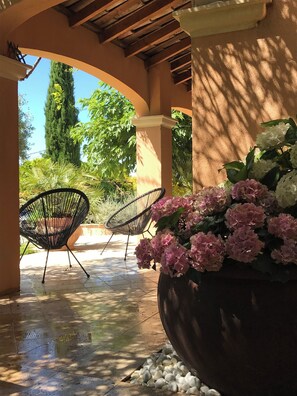 An outdoor patio area with a tiled floor, two black wire chairs, a large planter with pink and white flowers, and a wooden pergola structure. - Villa hauteur de Lauris infinity pool not overlooked (Lauris)