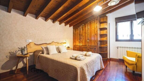 Um quarto com teto de madeira, uma cama com cabeceira, mesa de cabeceira com abajur, guarda-roupa e uma poltrona amarela.