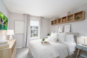 Ein Schlafzimmer mit einem Bett, Nachttischen, einem Fenster mit Vorhängen, einem an der Wand befestigten Regal mit Büchern und einer Pflanze sowie einem gerahmten Bild.