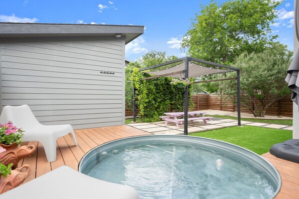 A backyard with a hot tub, a wooden deck, a white chair, a picnic table, and a pergola with string lights.