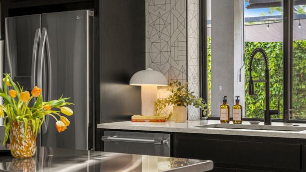 Eine moderne Küche mit einem Edelstahl-Kühlschrank, einer Vase mit Tulpen, einer Lampe und einem Fenster mit Grünpflanzen außen.