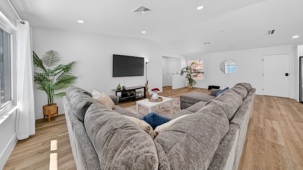 A spacious living room with a large grey sofa, a flat-screen TV mounted on the wall, and a potted plant.