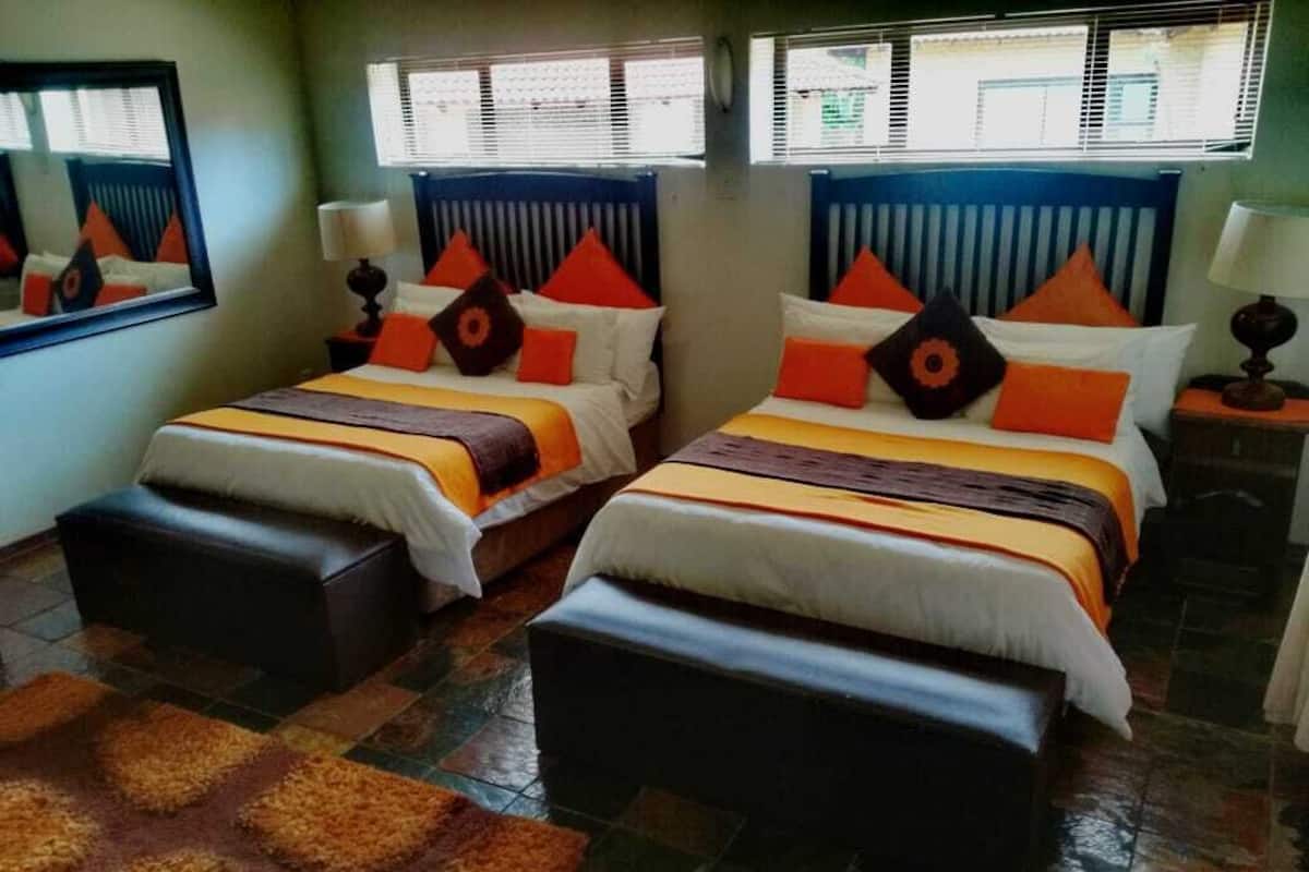 Una habitación con dos camas, cada una con cabecero, una mesita de noche con lámpara y un banco.