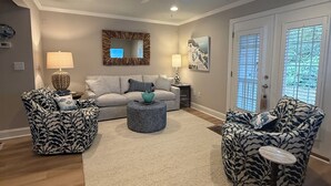 Un salon comprenant un canapé, deux fauteuils à motifs, une table basse, un ventilateur de plafond et une grande fenêtre avec des stores.
