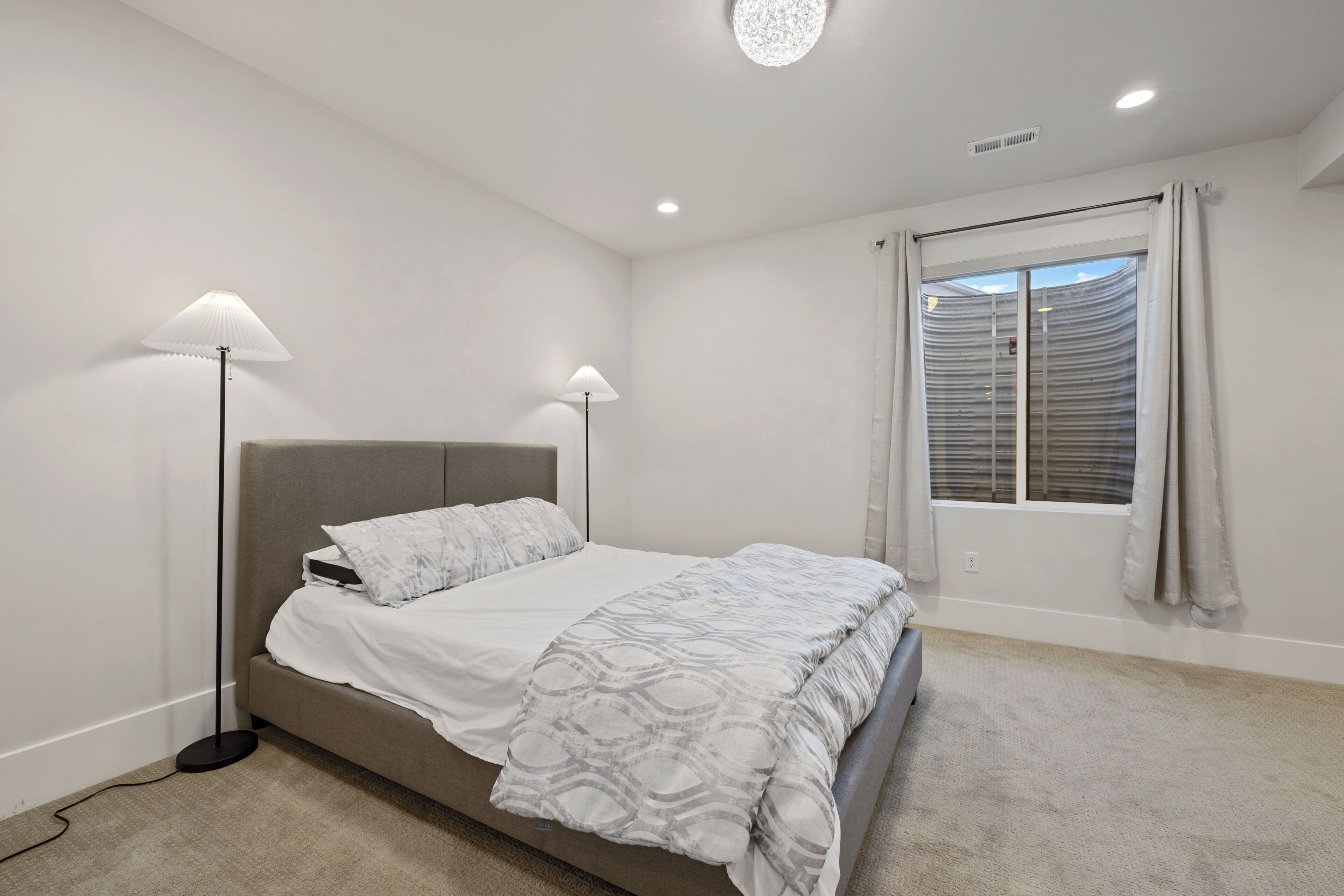 Ein Schlafzimmer mit einem Bett, zwei Stehlampen, einem Fenster mit Jalousien und einem Teppichboden.