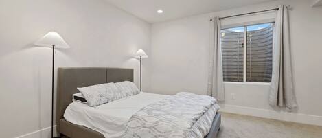Ein Schlafzimmer mit einem Bett, zwei Stehlampen, einem Fenster mit Jalousien und einem Teppichboden.