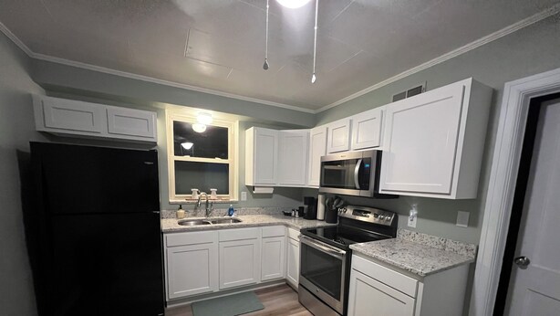 A compact kitchen with white cabinets, a black refrigerator, stainless steel appliances, and a wooden floor.