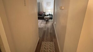 A narrow hallway with a patterned rug leading to a living area with a couch, a side table, and a lamp.