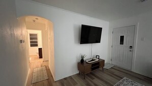 A hallway with a wooden floor, a TV mounted on the wall, a door, and a small cabinet with a plant.
