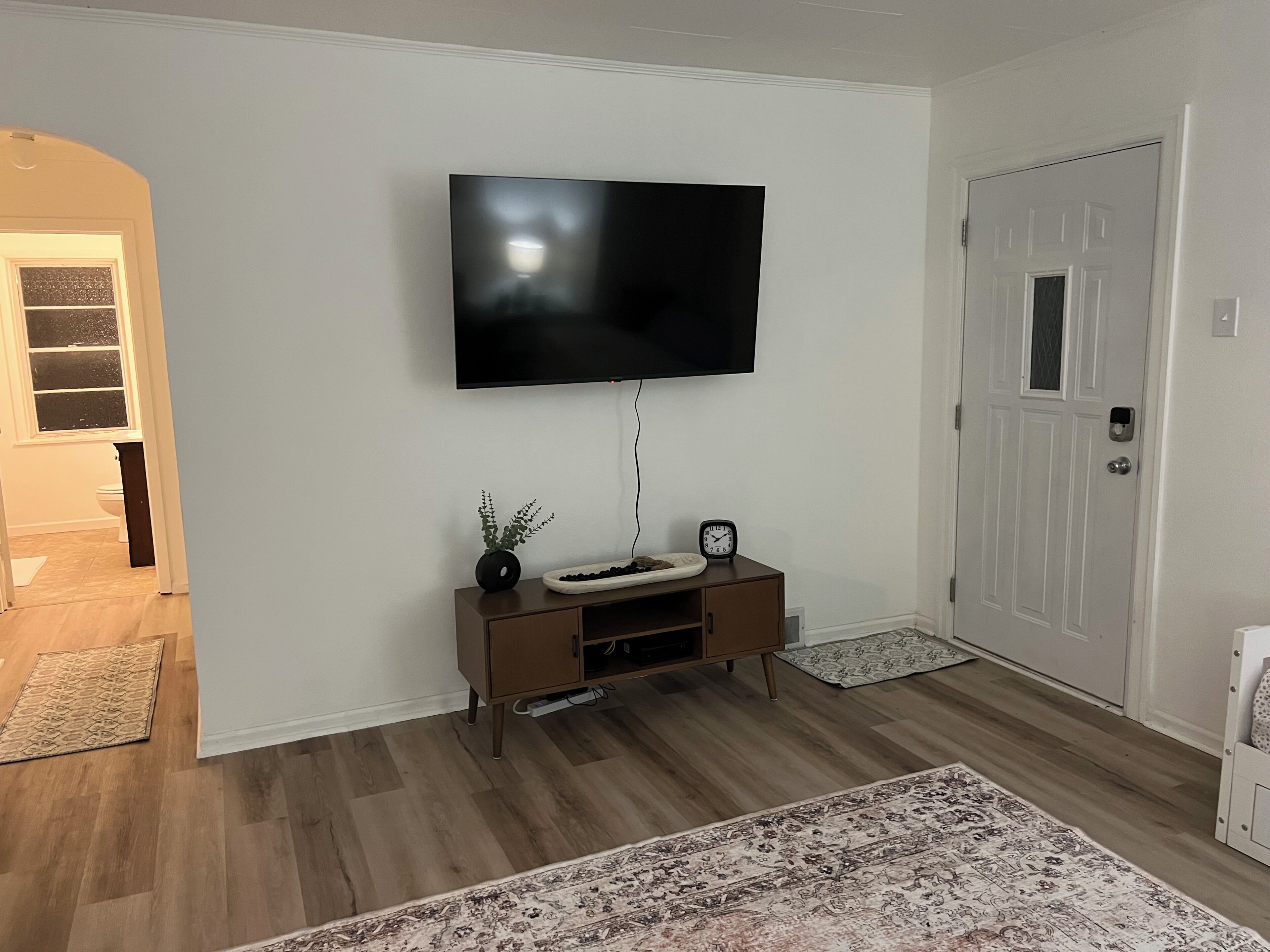 Un salón con un televisor de pantalla plana montado en la pared, un mueble marrón para entretenimiento con un reloj, y una alfombra estampada en el suelo.