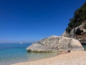 Un cielo azzurro limpido, un litorale roccioso e una persona sdraiata sulla spiaggia di ciottoli.