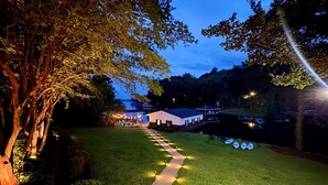 A nighttime view of a well-lit pathway leading to a building with a white exterior, surrounded by trees and a grassy area. - 8 bdrm covered dock, bar and kitchen on the water! hot tub, firepit, sleeps 26, (Henrico)