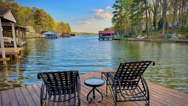 A wooden deck with two chairs and a small table, overlooking a calm lake with docked boats and a cabin in the background. - 8 bdrm covered dock, bar and kitchen on the water! hot tub, firepit, sleeps 26, (Henrico)