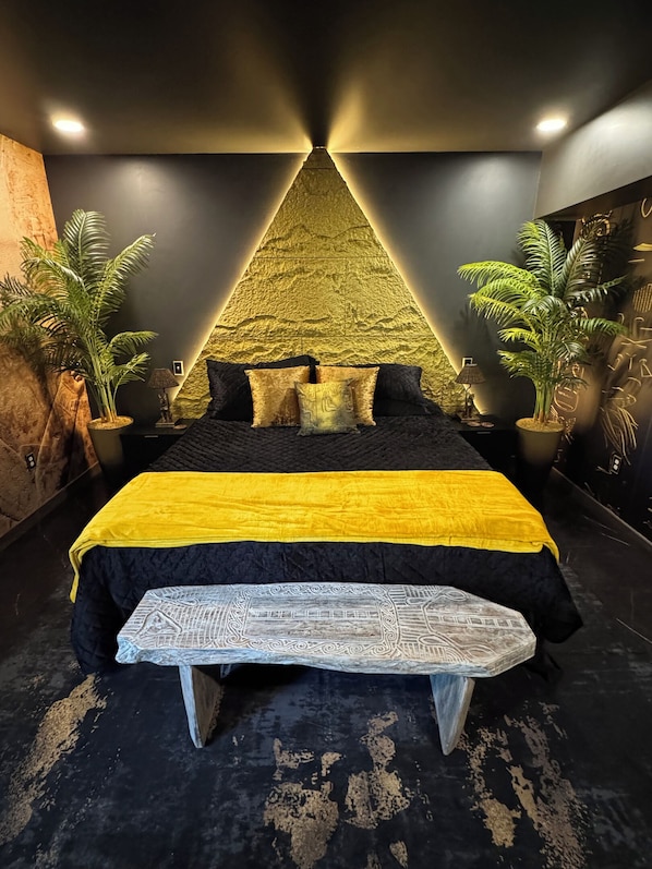 A bed with a yellow and black bedspread, a bench, and two potted plants.