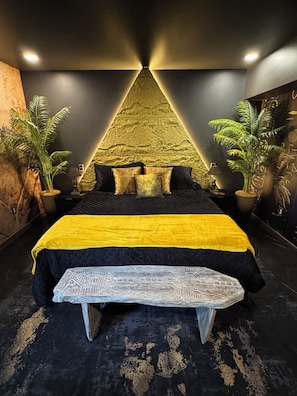 A bed with a yellow and black bedspread, a bench, and two potted plants.