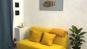 Un canapé jaune avec deux coussins, une œuvre d’art représentant un poisson encadrée au mur, une plante en pot et un tapis gris sur le sol.