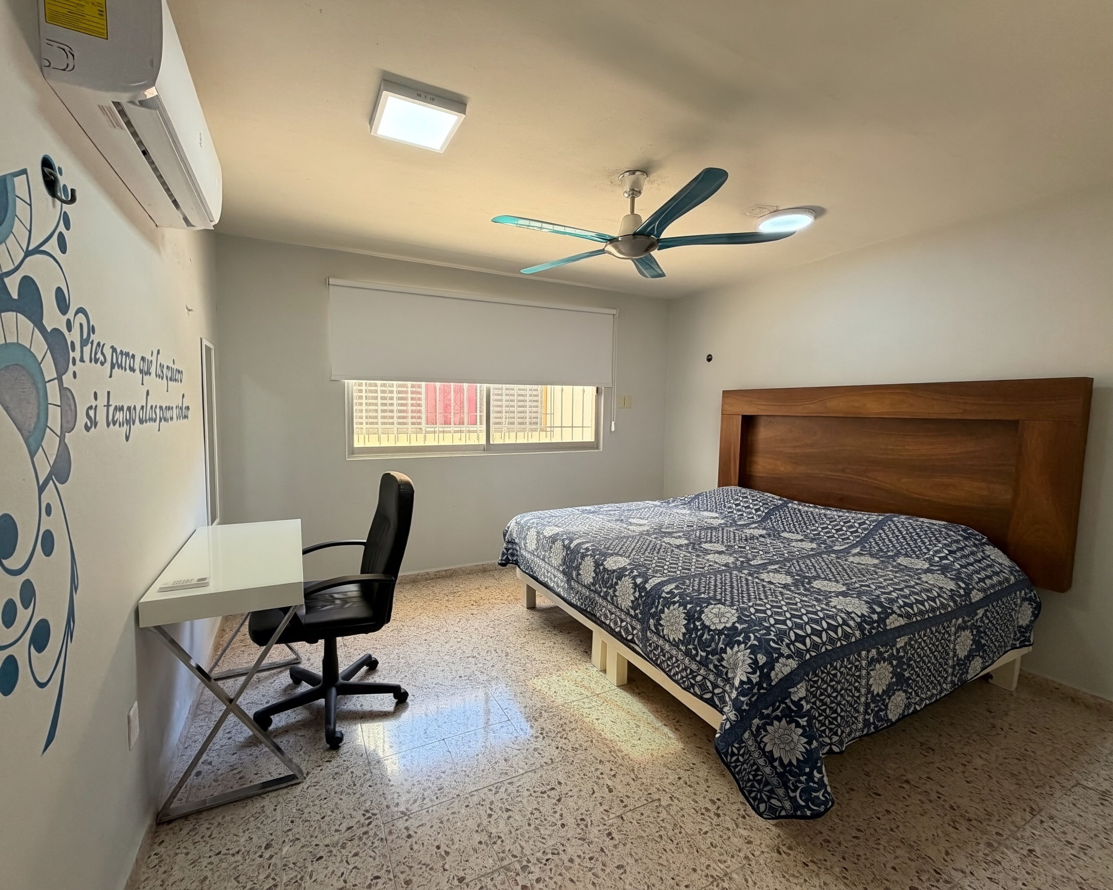 Une chambre à coucher comprenant un lit, un bureau, une chaise, un ventilateur de plafond et une fenêtre.