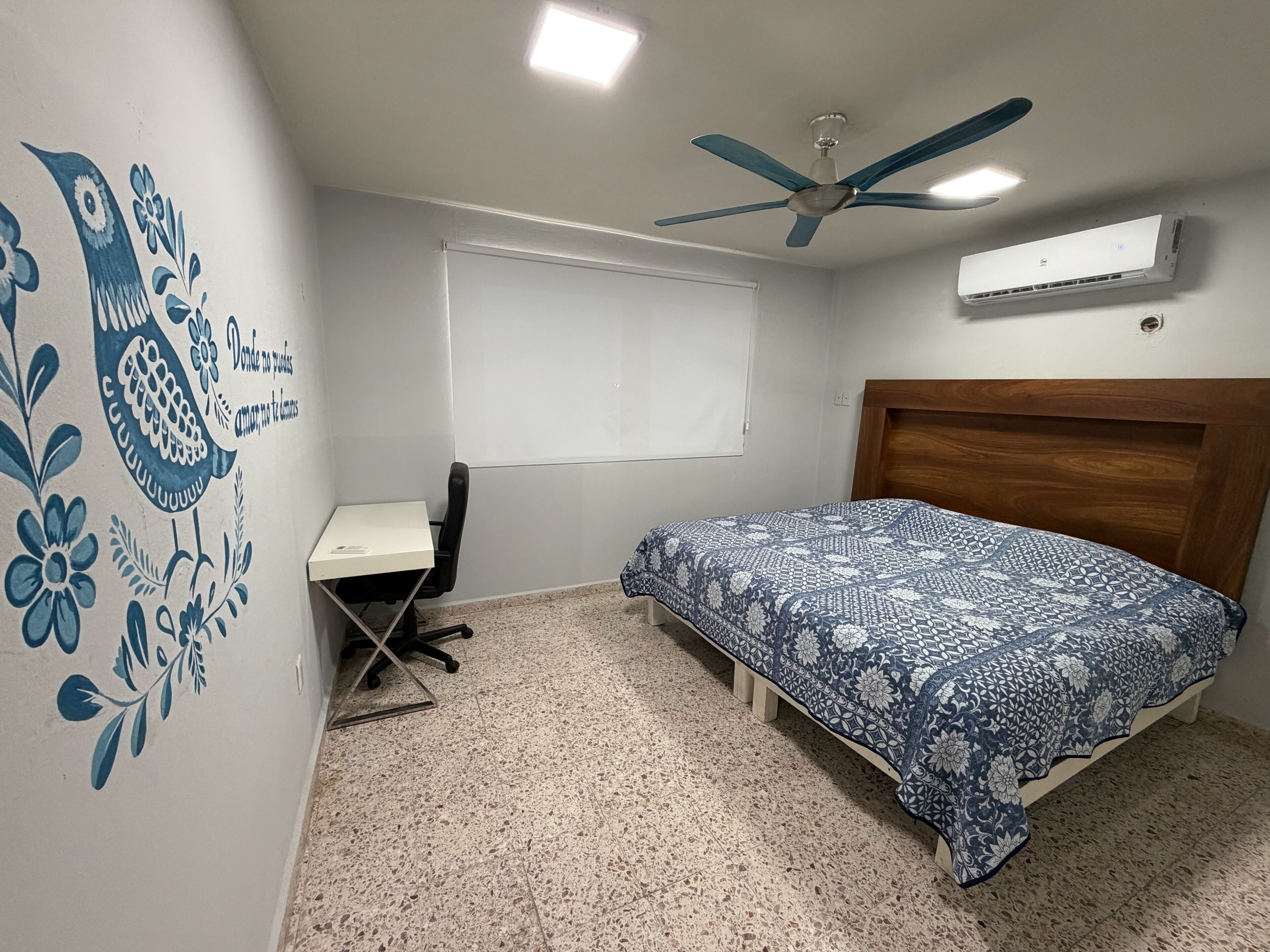 A bedroom with a bed, a desk, a chair, a ceiling fan, an air conditioner, and a wall with a floral design.