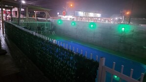 A nighttime view of a pool area with a white fence, green plants, and a building with a sign in the background. - Pousada Ilha do Itacolomi (Penha)