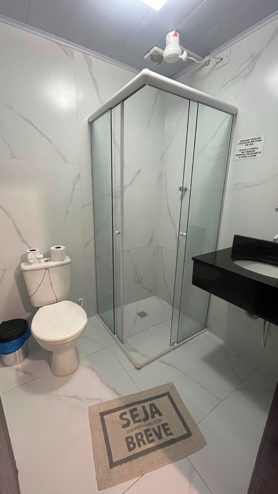 Un baño con ducha envuelta en vidrio, inodoro, lavamanos y felpudo con texto.