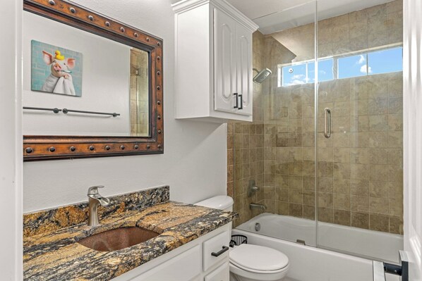 A bathroom with a granite sink, a toilet, a shower with a glass door, and a mirror with a decorative frame.