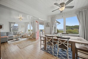A spacious living area with a sofa, coffee table, and dining table with chairs. There is a ceiling fan, large windows with curtains, and a view of the outdoors. - Spacious Group Getaway w/ Main House + Private Studio, Sleeps 10, Ideal for Families (Rockaway Beach)