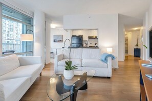 Ein modernes Wohnzimmer mit einem Glastisch, weißen Sofas und Blick zur Küche.