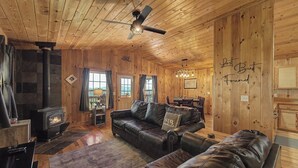 Interior de una cabaña de madera con chimenea, sofá de cuero, zona de comedor y ventilador de techo.