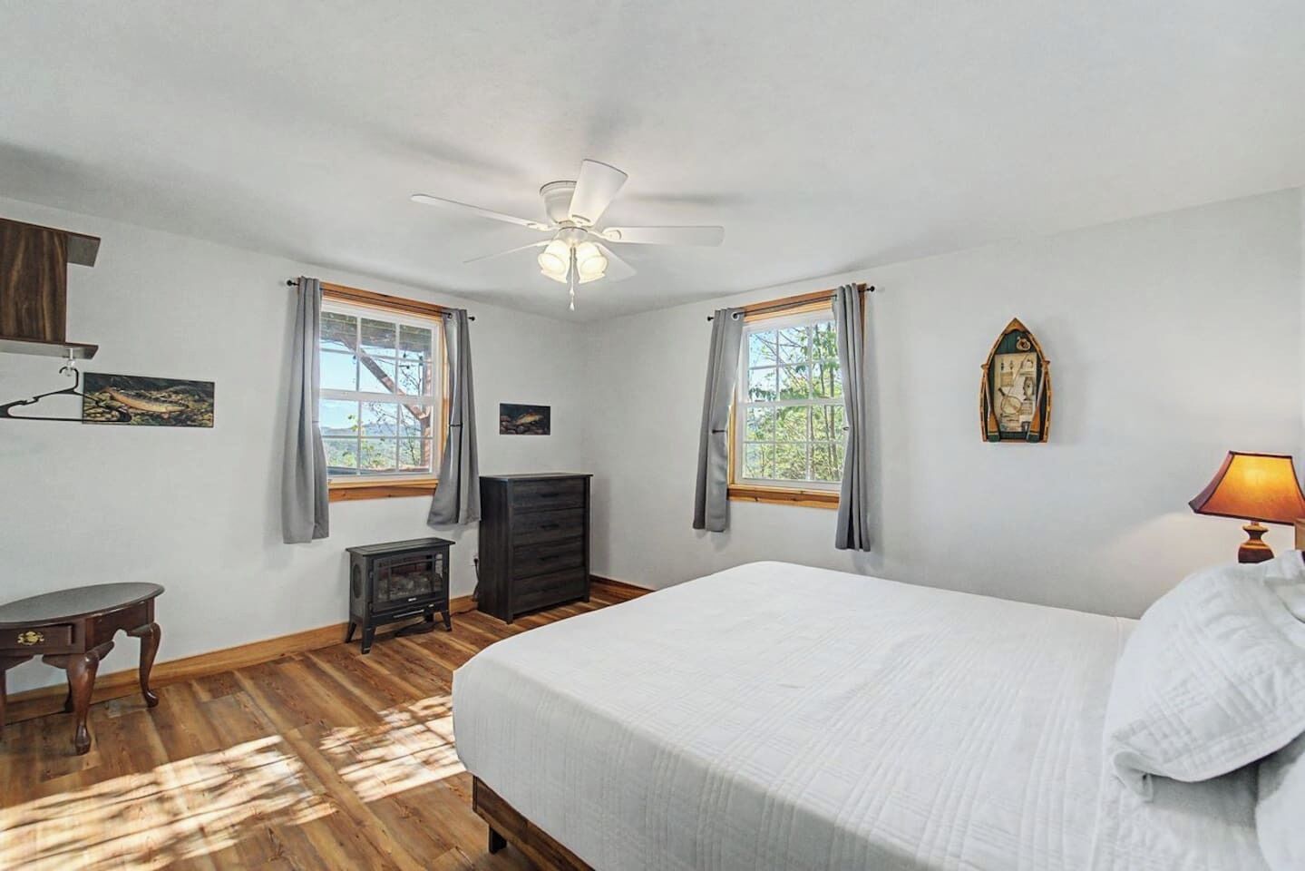 A bedroom with a bed, a ceiling fan, two windows with curtains, a dresser, a small table, and a lamp.