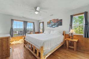 A bedroom with a wooden bed, a ceiling fan, a painting, and large windows.
