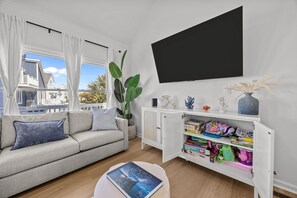 A living room with a sofa, a coffee table with a book, a TV, a cabinet with children's toys, and a large window with curtains.
