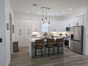 A modern kitchen with white cabinetry, a central island with bar stools, a stainless steel refrigerator, and pendant lighting. - THE TOGETHER PLACE 2 (naples)