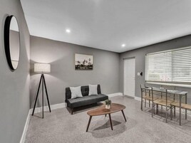 A modern living room with a gray sofa, a round coffee table, a floor lamp, and a dining area with a metal table and chairs.