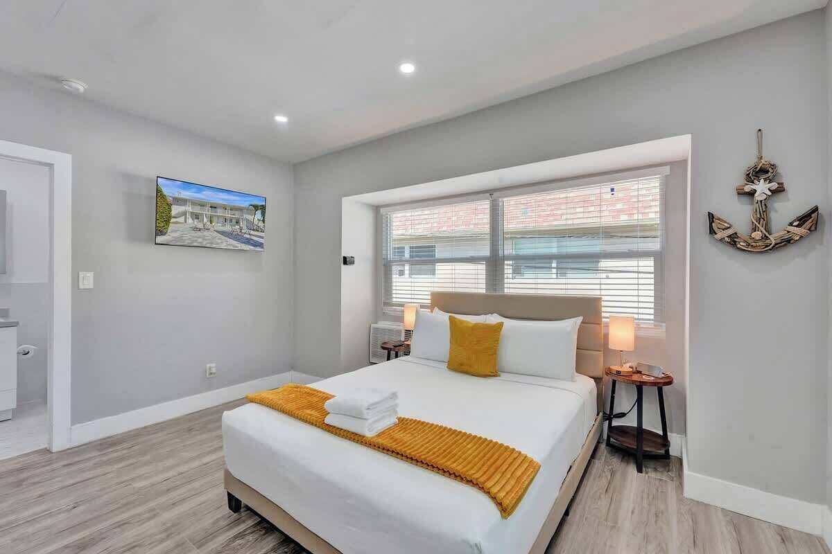 A modern hotel room with a large bed, a window with blinds, a small table with a lamp, and a wall-mounted decorative anchor.