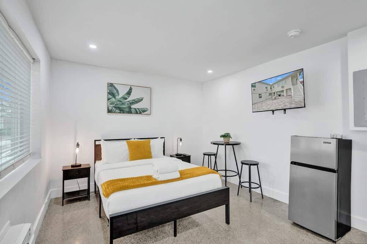 A modern hotel room with a bed, a desk, a chair, a refrigerator, and a window.