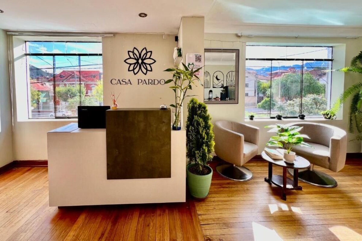 A reception desk with a logo, a potted plant, a sofa, and a view of buildings through large windows.