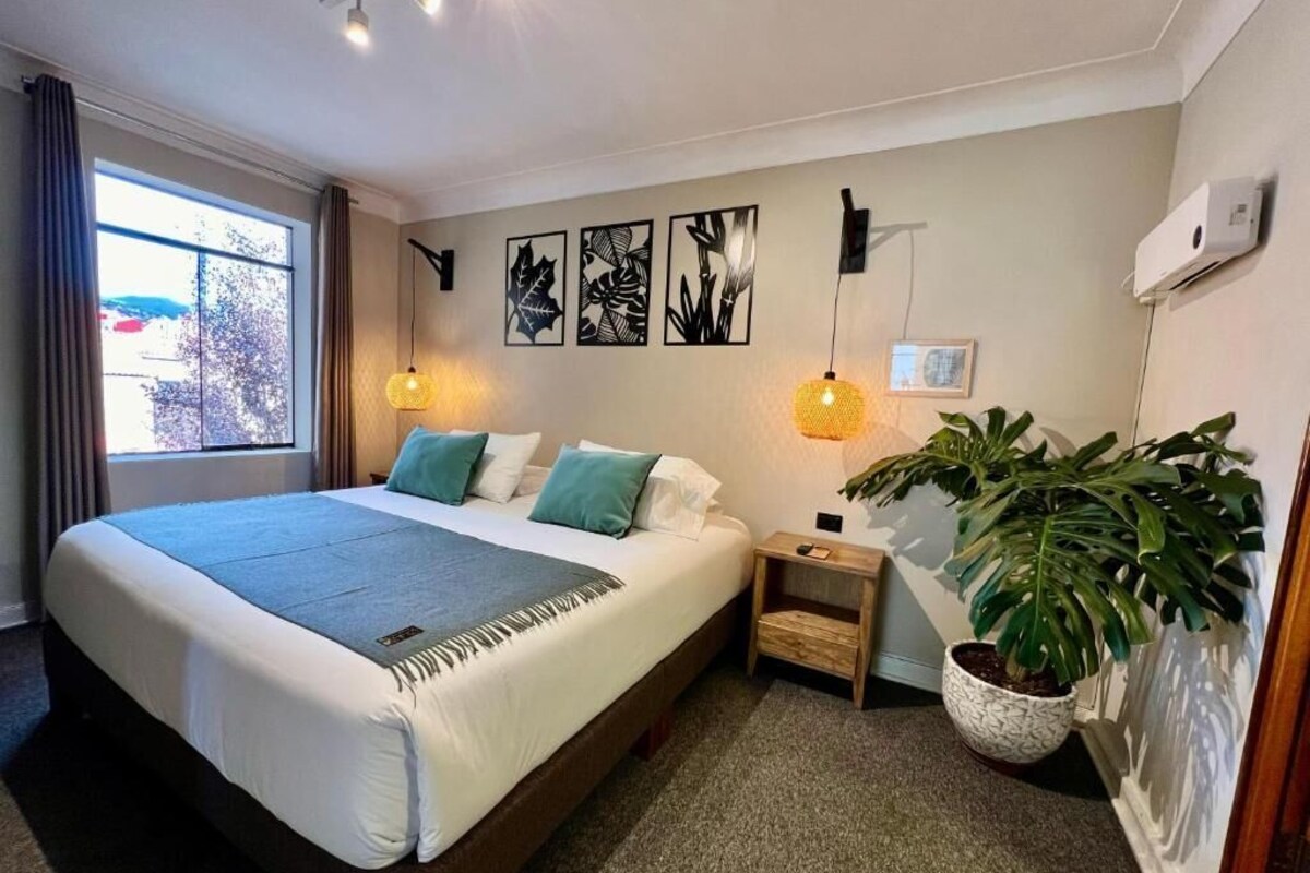 A bedroom with a large bed, bedside tables, a potted plant, wall art, and a window with curtains.