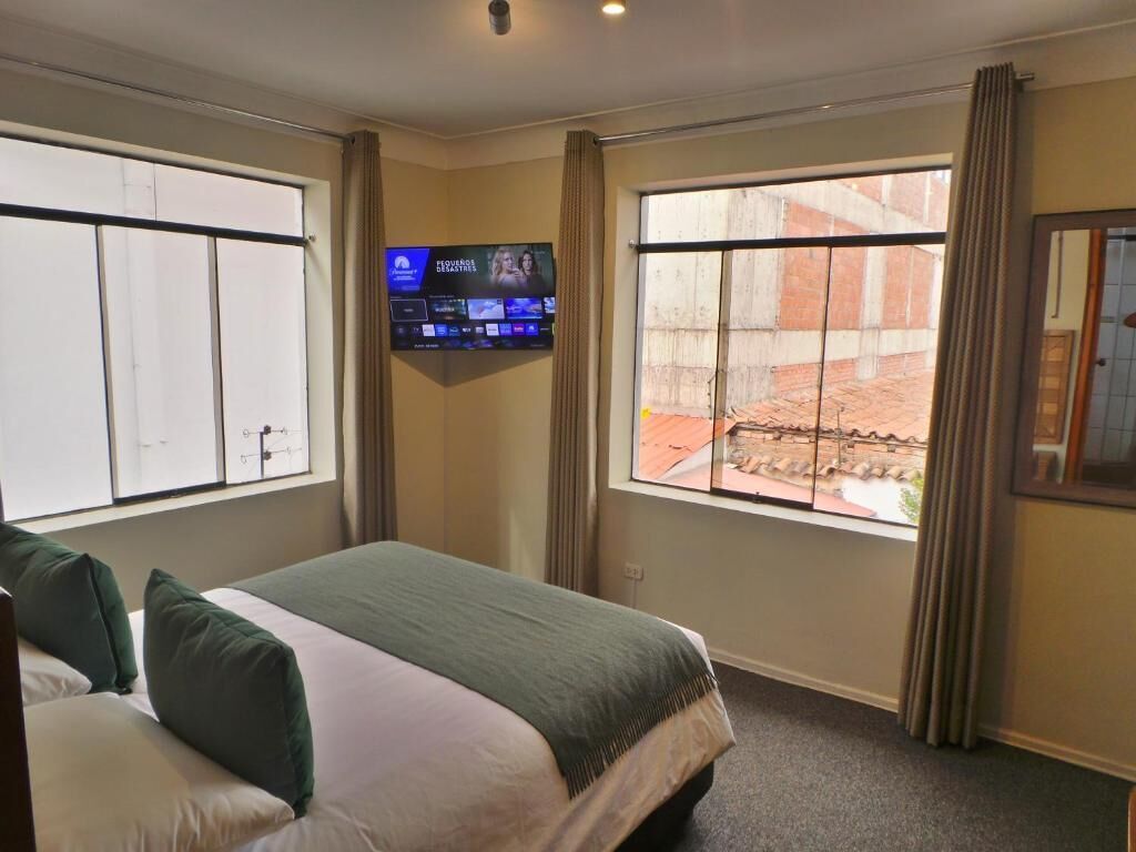 Ein Hotelzimmer mit zwei Betten, einem an der Wand montierten Fernseher und großen Fenstern mit Vorhängen.
