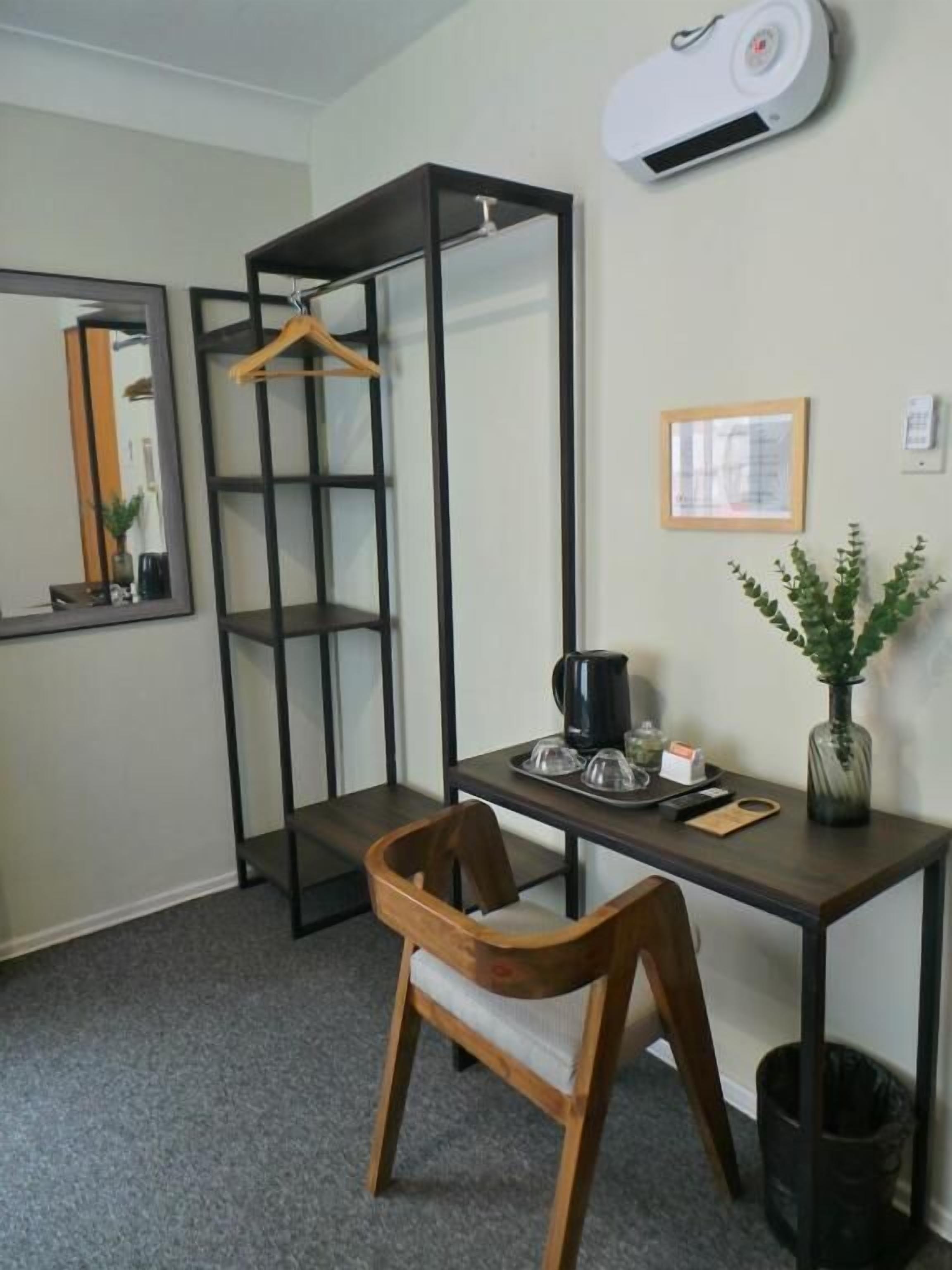 A room with a desk, chair, hanging clothes rack, mirror, framed picture, and a potted plant.
