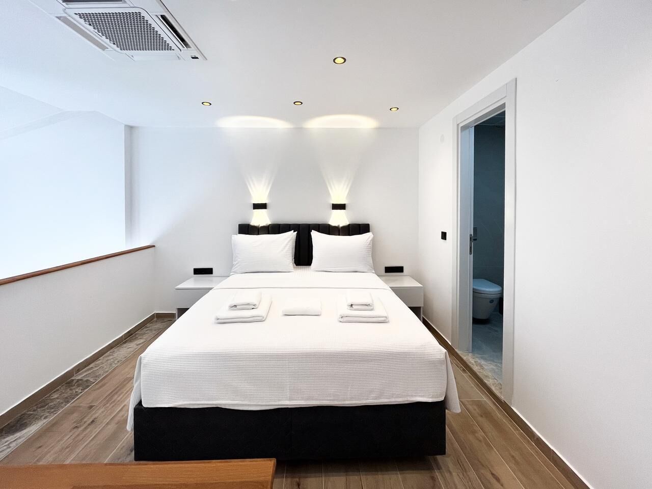 A modern hotel room with a large bed, bedside tables, a wooden floor, and a view of the hallway.