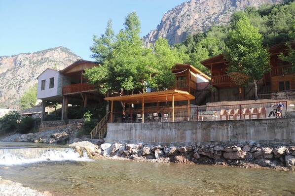 A riverside hotel with outdoor seating, a stone retaining wall, and mountainous background. - Kapuzbasi Uçar Pansiyon (Yahyali)