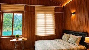 Ein Schlafzimmer mit Holzbalkendecke, einem Bett mit gemusterten Textilien, einem Nachttisch, einer Lampe, einem kleinen Tisch mit Pflanze und einem Fenster mit Jalousien.