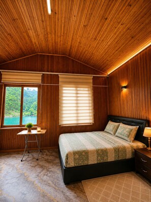 A bedroom with a wooden ceiling, a bed with patterned bedding, a nightstand, a lamp, a small table with a plant, and a window with blinds.