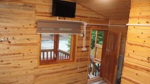 Ein Holzblockbau-Innenraum mit an der Wand montiertem Fernseher, einem Fenster mit Blick ins Freie und einer Tür zu einer Außenterrasse.