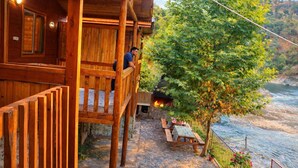 Eine Holzhütte mit einem Balkon mit Blick auf einen Fluss, einer gepflasterten Fläche mit Tisch und Stühlen sowie einer Feuerstelle.