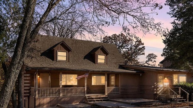 A two-story log cabin with a porch, a flag, and a staircase leading to the entrance. - Lakefront Home w/ Private Apartment | Sleeps 12 | Boat Slip Available (Branson West)