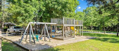 A playground with swings and slides, surrounded by trees and a clear sky.