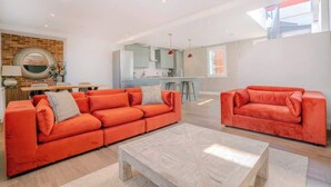 A modern living room with two orange sofas, a wooden coffee table, and a kitchen in the background. - Wimbledon 3-Bed Townhouse | Roof Terrace + Parking | Sleeps 6 (Greater London)