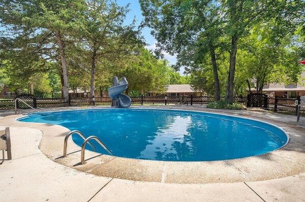 A swimming pool with a slide, surrounded by trees and a fence.
