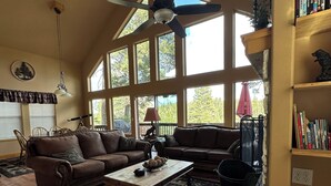 Una sala de estar amplia con techo alto, ventanas grandes, ventilador de techo, chimenea, estantería y comedor.